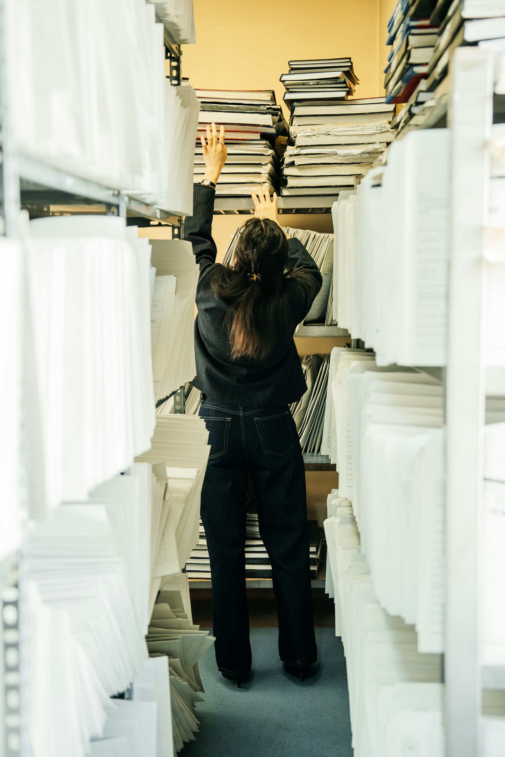 A young woman reaching for a book on a high shelf.