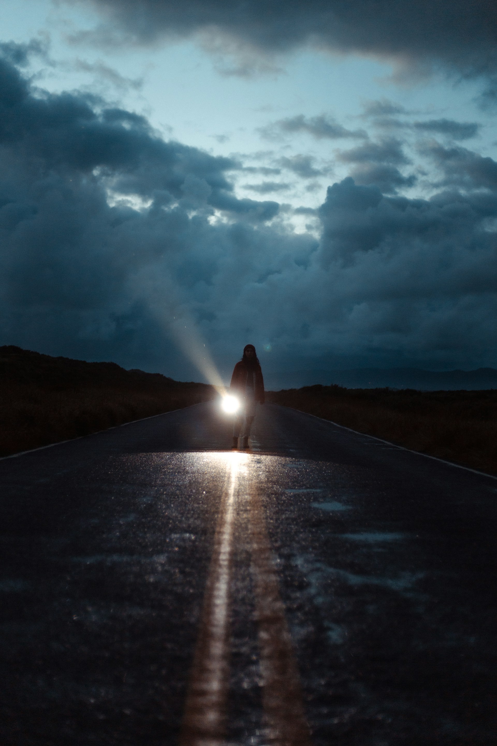 A person standing alone on a dark road holding a powerful flashlight.